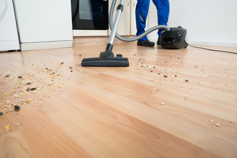 A professional from Mr Sander® vacuuming debris from a wooden floor in a kitchen area. The image shows the professional using a black canister vacuum to clean up various particles and ensure the floor is spotless before sanding and refinishing.
