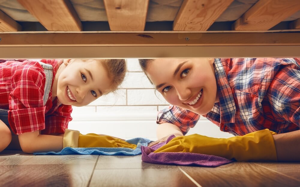 An adult and a young child wearing protective gloves, smiling while cleaning a hardwood floor under a bed.