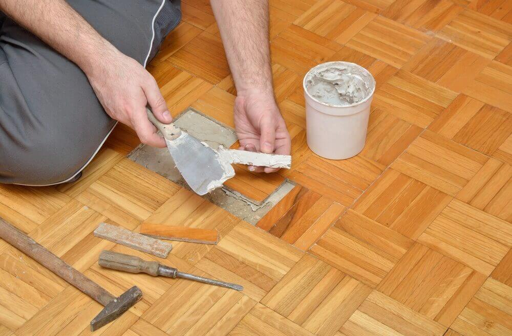 A person is repairing a wooden parquet floor using a putty knife to apply adhesive or filler in a damaged area. A container of filler is nearby, along with other tools such as a hammer and chisel, indicating a floor restoration in progress.