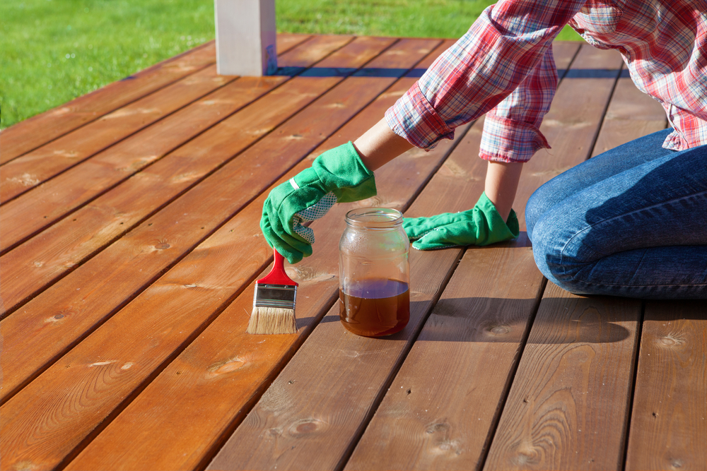 Person in a plaid shirt and green gloves staining a wooden deck with a brush and jar of stain.