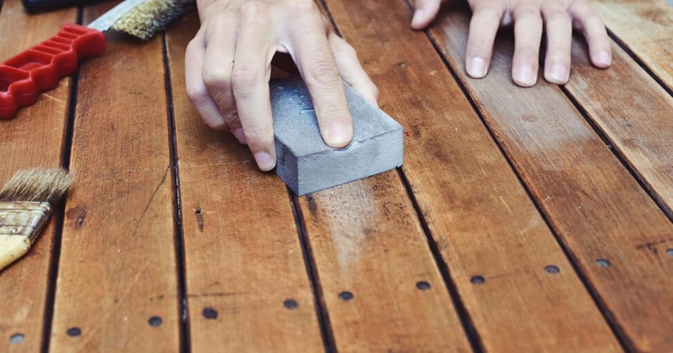 A person's hand holding a sanding block on a wooden plank surface, with a paintbrush and wire brush lying nearby.