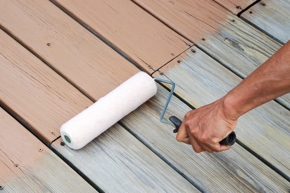 Hand with a paint roller applying sealant to wooden deck boards.