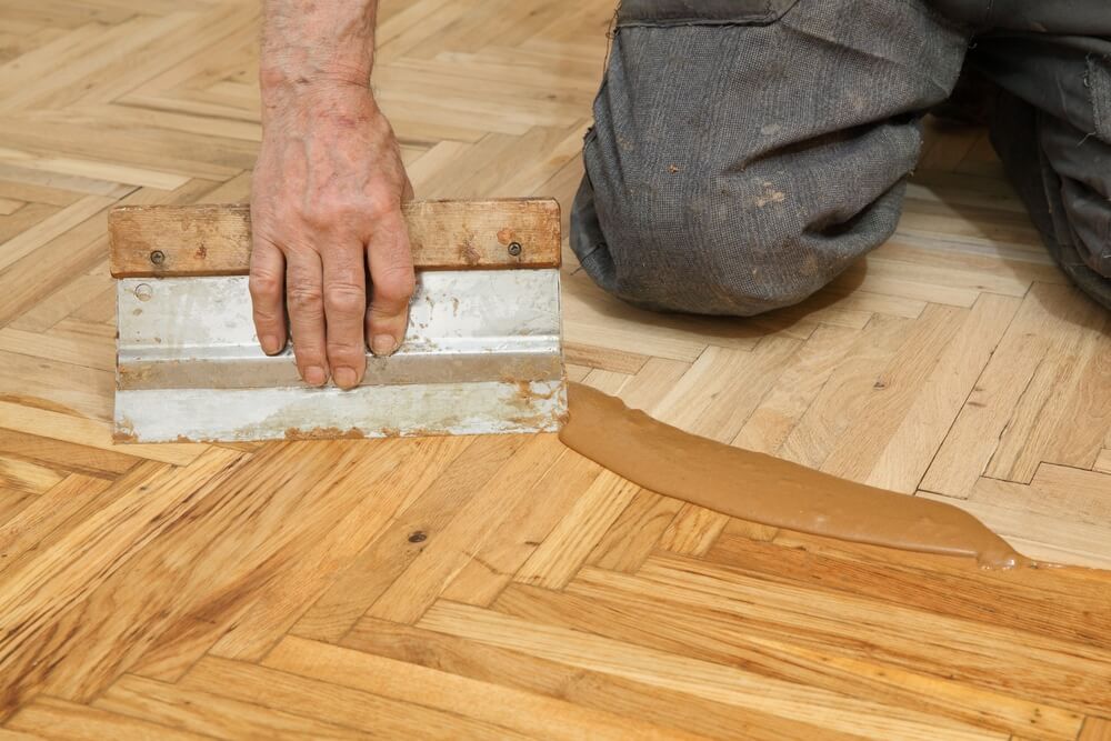 A professional from Mr Sander® kneeling and using a trowel to apply Bona Pacific Filler to a herringbone parquet floor during a sanding and refinishing project in a UK living room. The filler is spread evenly to fill gaps and cracks in the wood, preparing the floor for a smooth finish.