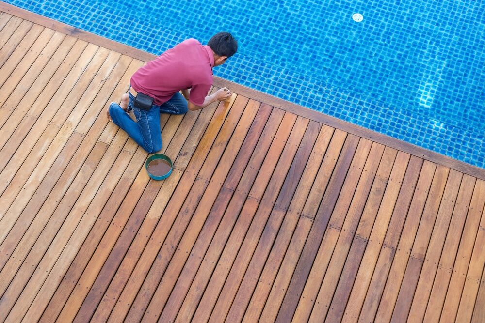 A person staining a wooden deck beside a swimming pool.