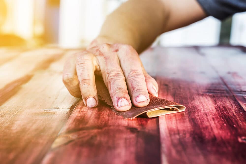 Close-up of a hand sanding a wooden surface with a piece of sandpaper.