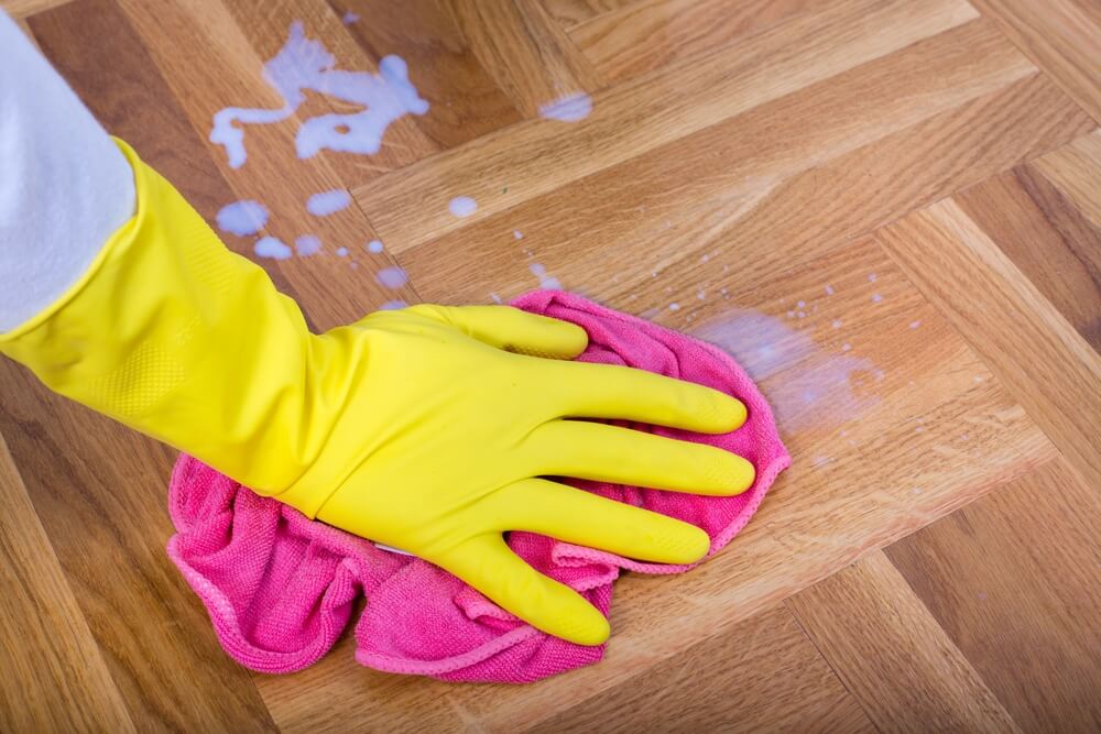 A close-up view of a person wearing yellow rubber gloves cleaning a wooden parquet floor with a pink cloth and cleaning solution. The floor has a herringbone pattern, and the cleaning solution is visibly spread on the surface.