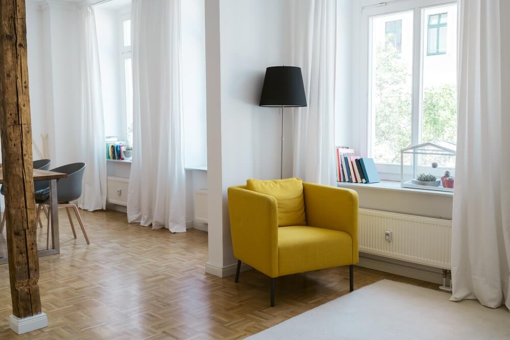 A bright and airy living space featuring a newly refinished parquet floor, bathed in natural light from large windows. The room is tastefully decorated with white curtains, a vibrant yellow armchair, and a sleek black floor lamp. A dining area with modern chairs and a rustic wooden pillar adds charm and character to the room.