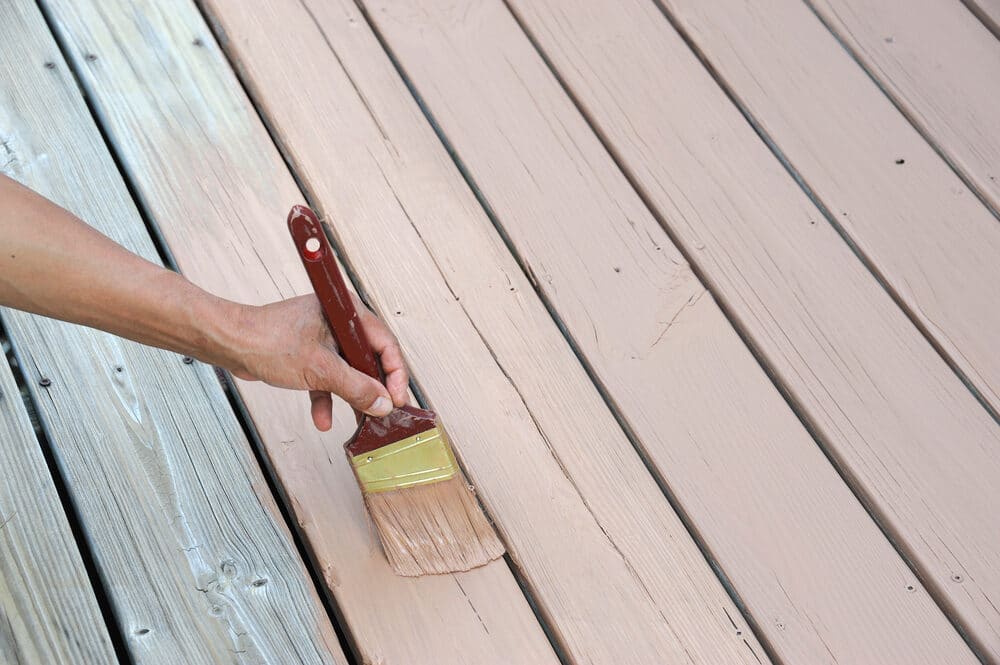 A person’s hand holding a paintbrush, applying light brown paint to a wooden deck.