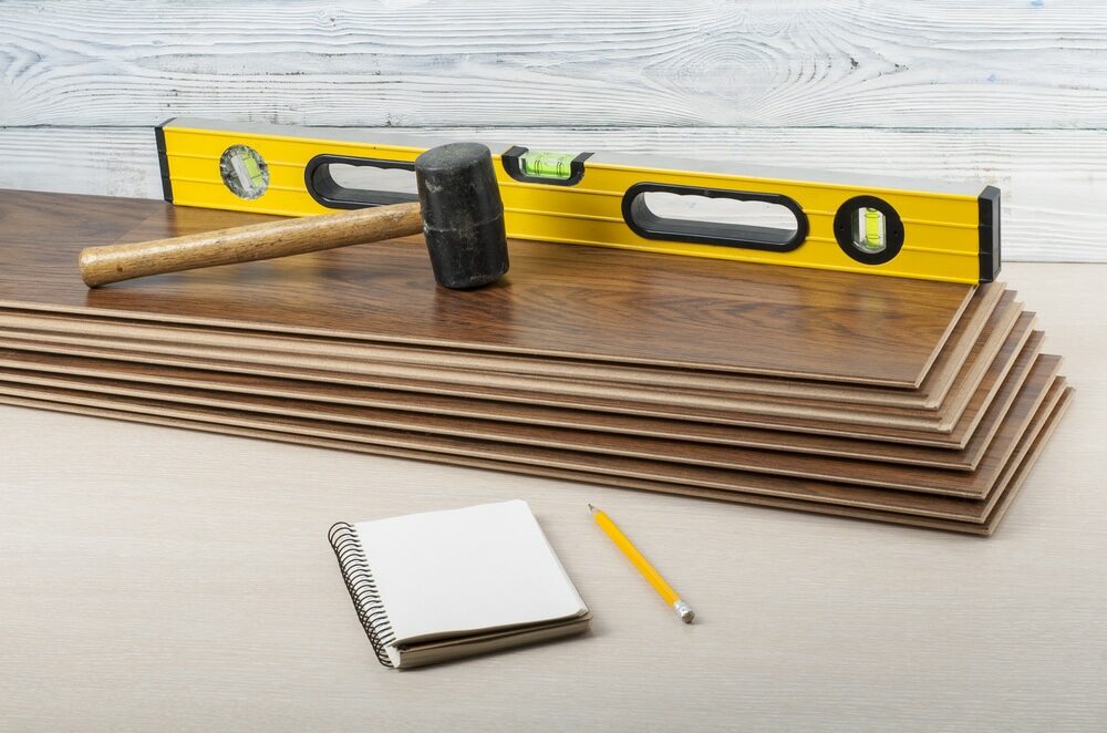 A spirit level, rubber mallet, notebook, and pencil on top of laminate flooring planks.