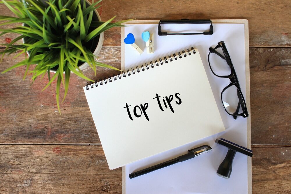 A flat lay photograph of a desk with a spiral notebook opened to a page with "top tips" written on it. Next to the notebook is a pair of black glasses and a black pen, with a clipboard and decorative items like a potted green plant and a heart-shaped paper clip above the notebook. The wooden texture of the desk provides a warm background, creating a setting that suggests planning or brainstorming.