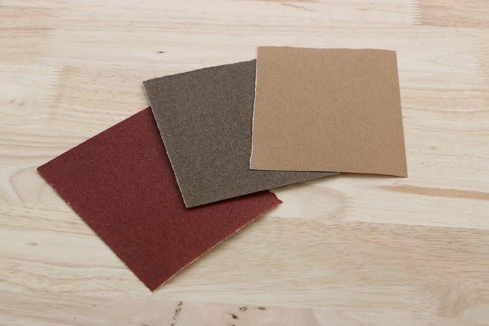 Three different grit sizes of sandpaper sheets on a wooden surface.