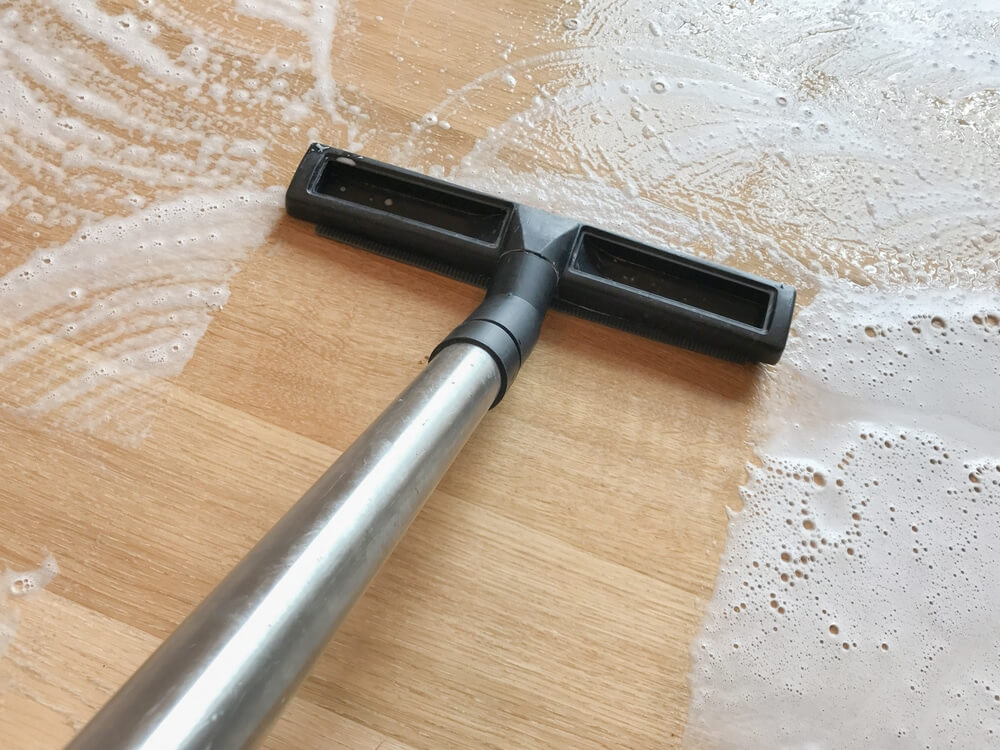 A close-up of a wooden floor being cleaned during the sanding and refinishing process by Mr Sander®. A vacuum head with a stainless steel handle is being used to remove soapy water from the floor. The surface is covered in foam, indicating thorough cleaning as part of the refinishing procedure.
