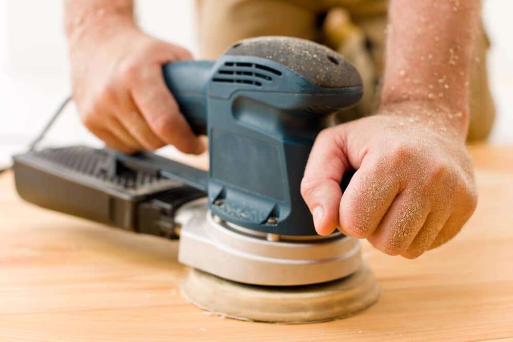 Close-up of a person using an electric sander on a wooden surface, with sawdust covering their hand.