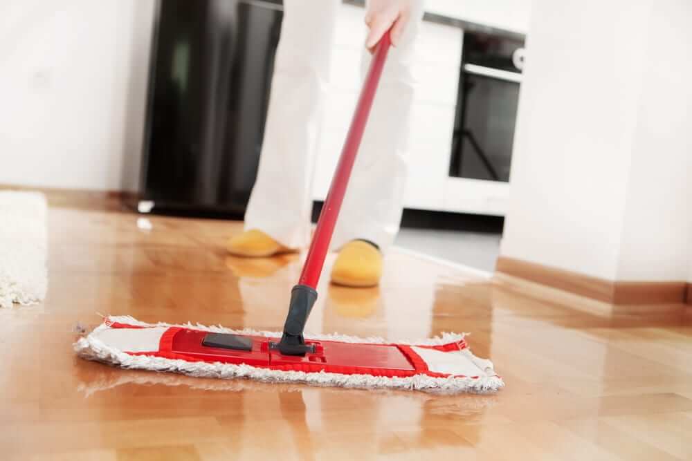 A person cleaning a newly refinished wooden floor using a red mop in a bright and modern living room. The floor has a high-gloss finish, reflecting the light, indicating a professional sanding and refinishing job by Mr Sander®. The scene includes a glimpse of kitchen appliances and a rug.