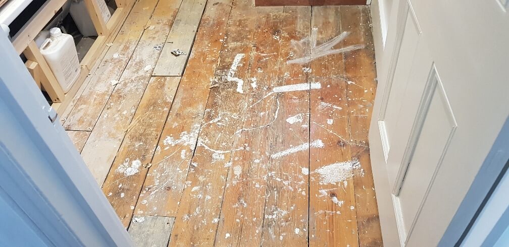 A wooden floor in a room showing heavy wear, paint splatters, and visible gaps between the floorboards. The floor appears to be in need of repair and refinishing, indicating a pending restoration project.
