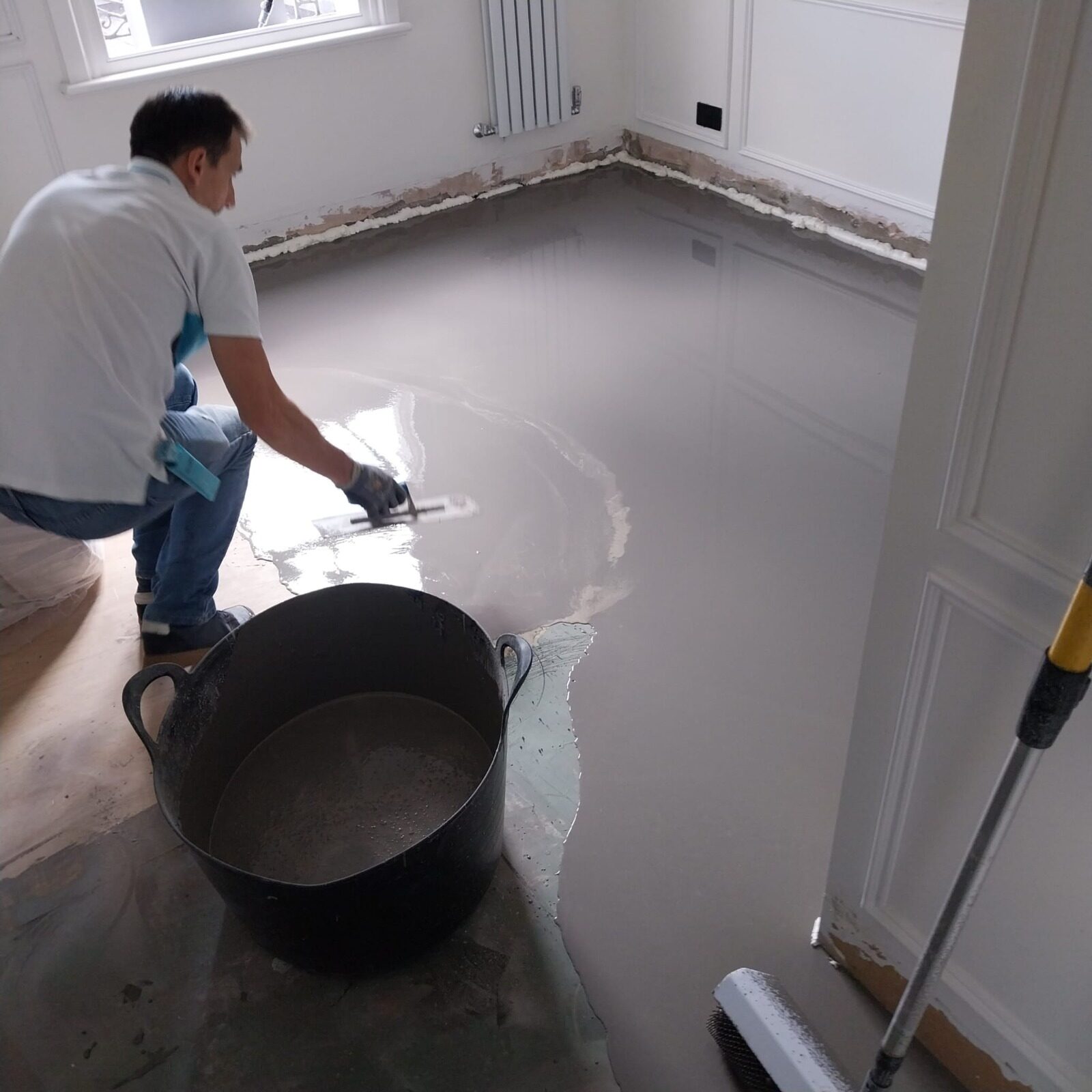 Technician applying self-levelling compound to prepare a screed for Solid Boards Over UFH, prior to fitting a solid oak hardwood floor (solid wood boards).