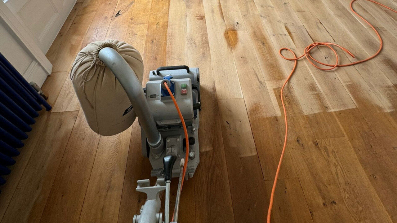 Belt sander working on solid oak hardwood plank flooring during professional Sanding and Finishing, showing partially sanded boards and dust-extraction bag