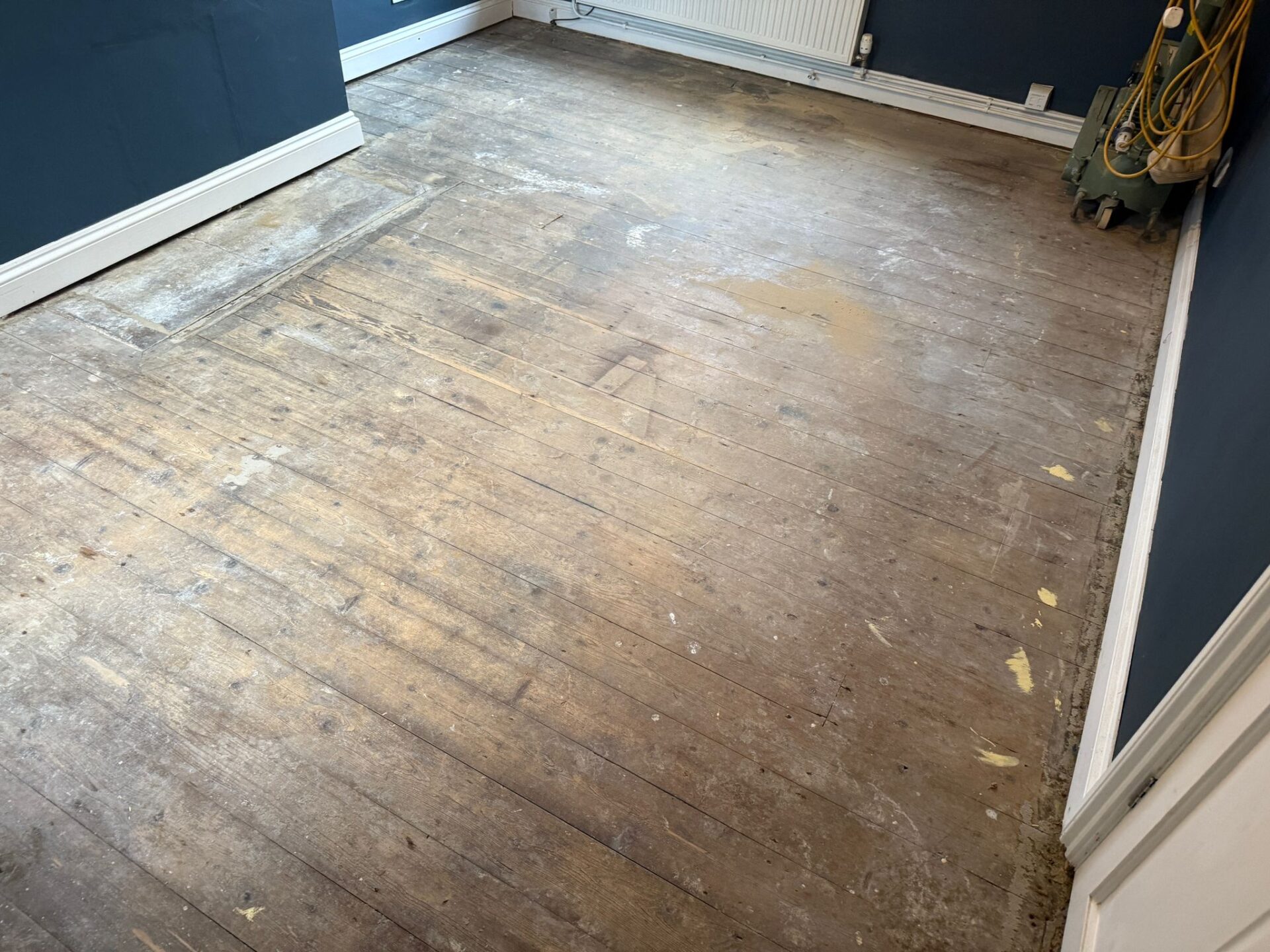 Worn original solid pine floorboards before Solid Wood Floor Sanding, showing scuffs, stains and patch repairs across the timber surface in a living space.