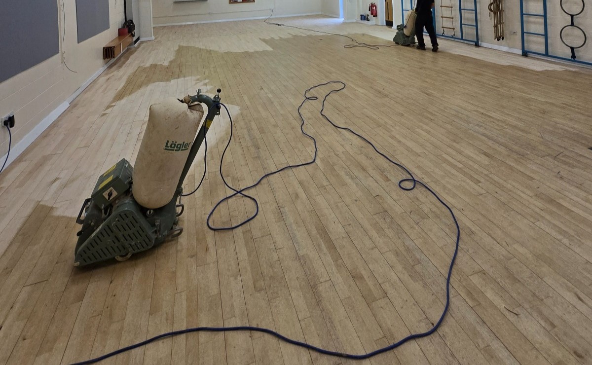 Sports Hall Floor Sanding in progress on a sprung maple strip sports hall floor, with belt sanders working across the worn hardwood surface.