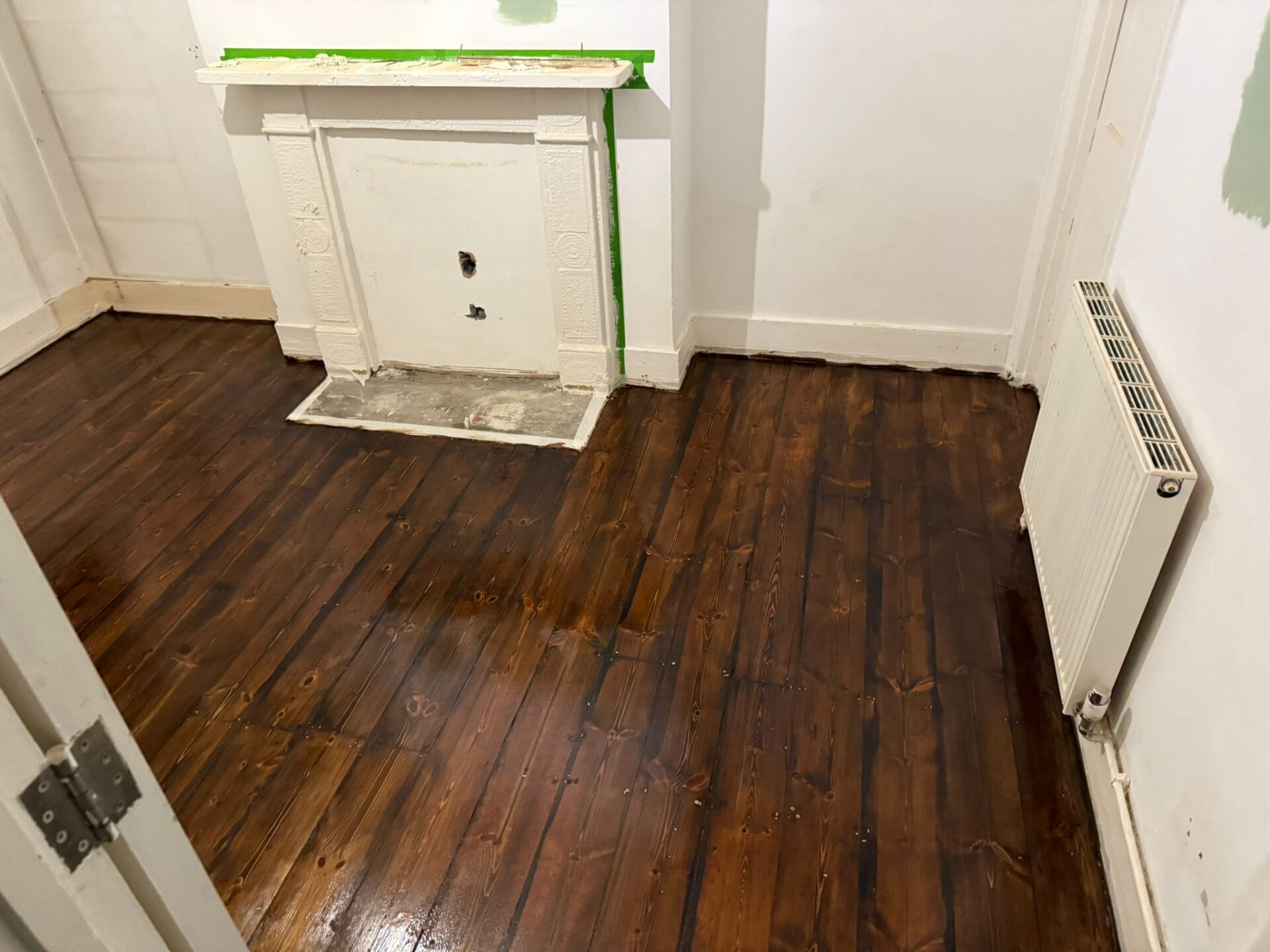 Freshly restored Victorian pine floorboards (solid softwood) after staining wood floors to a deep walnut tone; smooth, even colour across tongue-and-groove boards around a period fireplace and radiator.