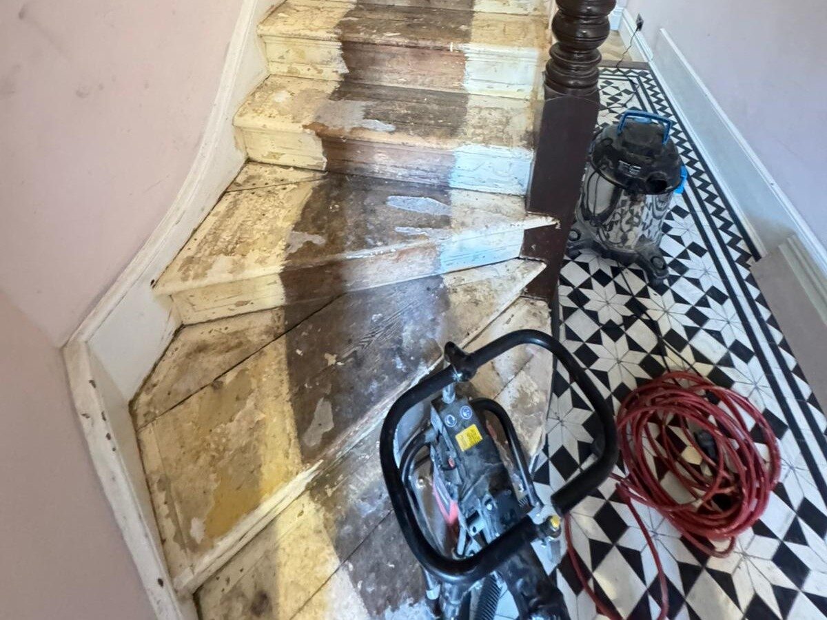 A staircase in progress showing the removal of old white paint and dark stain through the process of sanding stair treads on pine softwood.