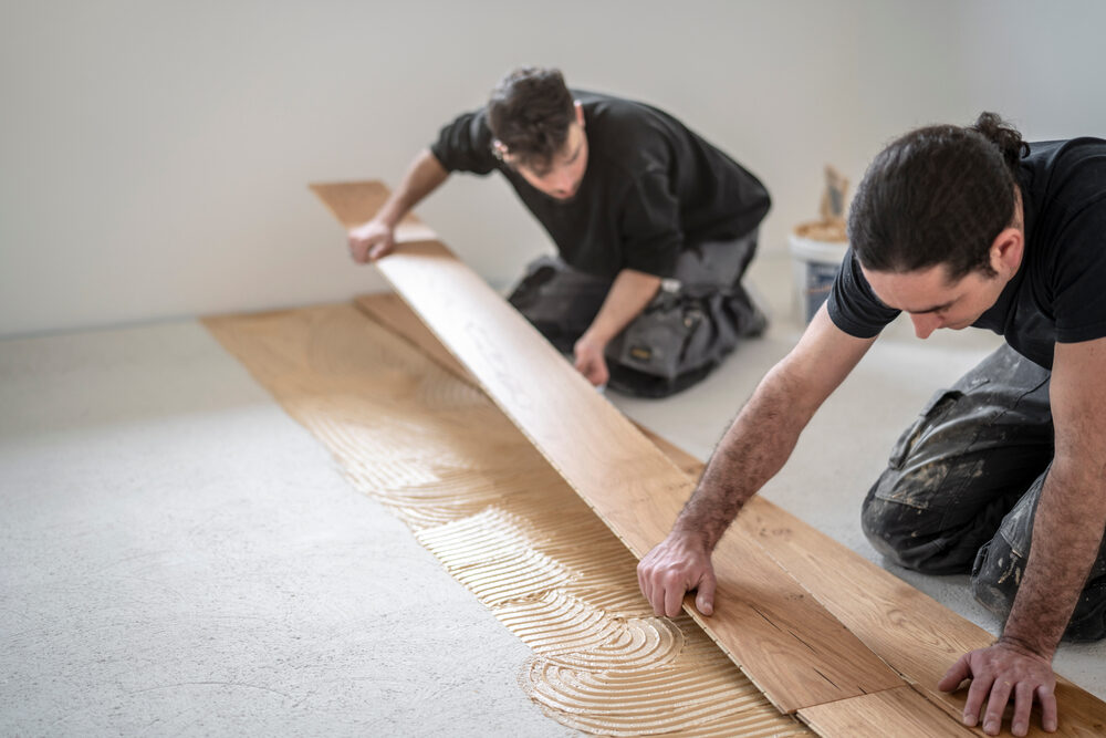 Two flooring specialists fitting a long light‑oak board onto fresh adhesive, creating an even wood floor surface for a photographic studio