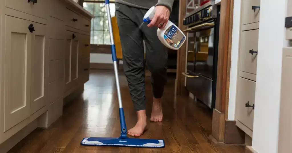 A person using a Bona floor cleaning product and a mop to clean dark wooden floors in a kitchen. The setting shows the lower half of the person, focusing on the mop and the cleaning product bottle they are holding. The dark wood floor shines, reflecting the light from the kitchen, indicating a clean and polished surface.
