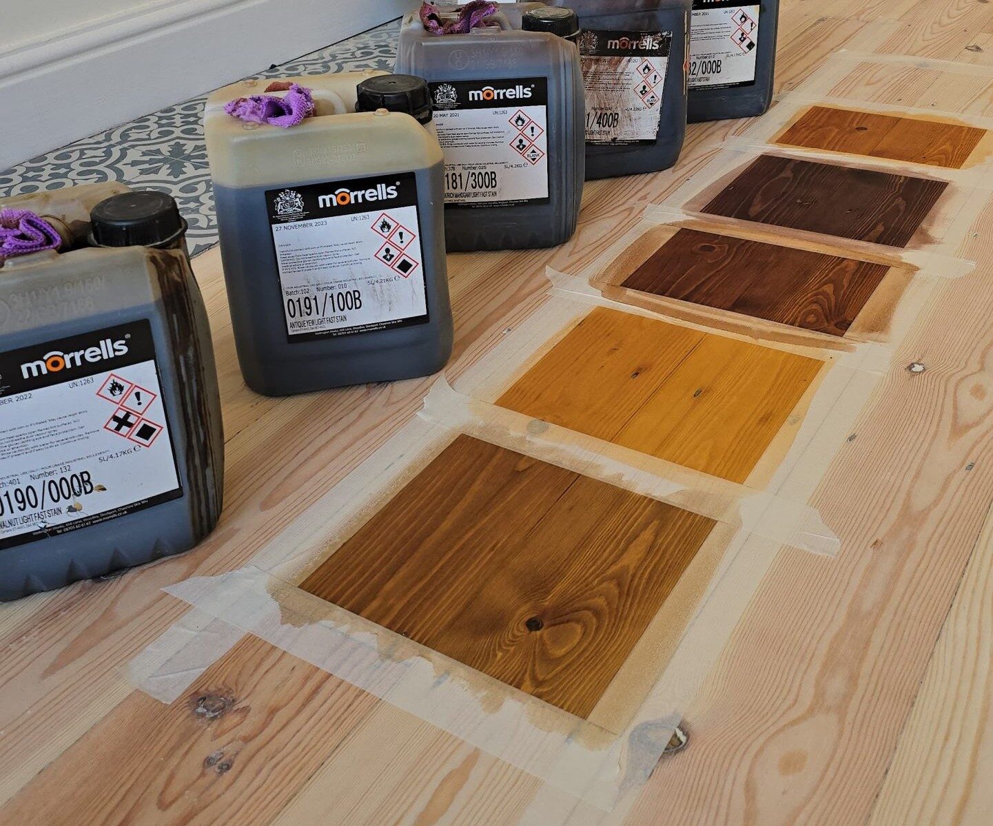 Stain sample test panels taped on sanded timber floorboards with finishing products, illustrating colour selection in The Lifecycle of Hardwood Floors.