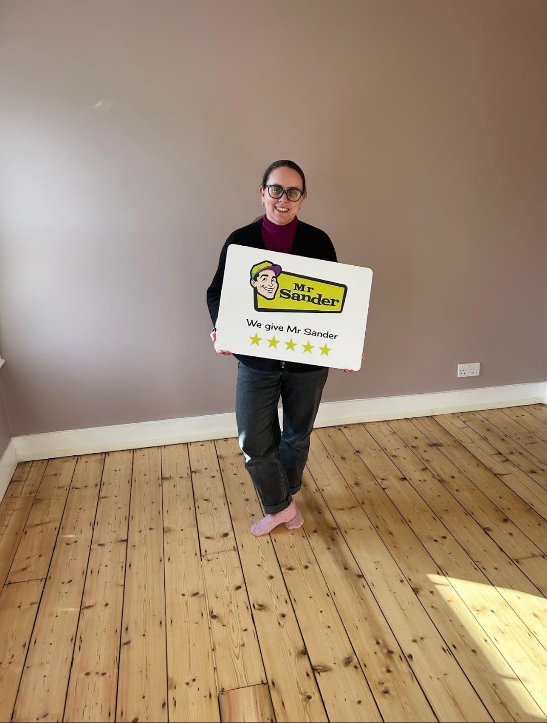 A happy customer standing on newly refinished wooden floors in Surrey, UK, holding a sign that reads "Mr Sander - We give Mr Sander 5 stars". The room has a warm, neutral wall color and plenty of natural light.