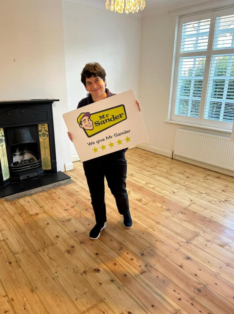 A satisfied customer holding a "Mr Sander" sign stands in a room with newly refinished wooden flooring, a black fireplace, white walls, and a window with white shutters.