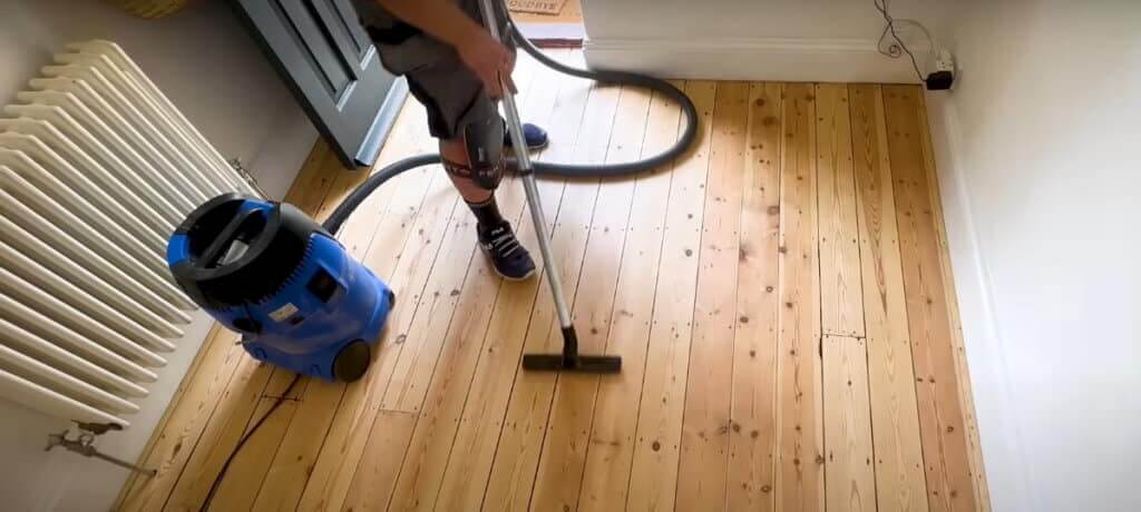 A person vacuuming a wooden floor using a blue vacuum cleaner in preparation for gap filling. The floor has visible gaps and knots, showing the need for cleaning before refinishing.