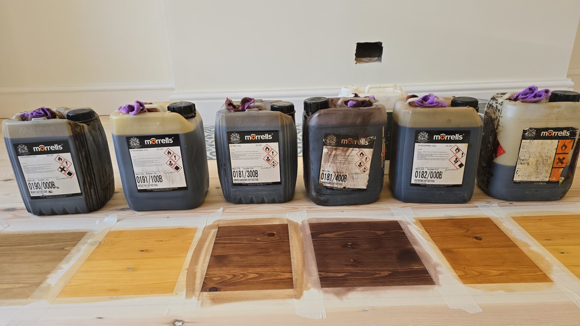 Victorian Pine Floor stain test—multiple colour samples brushed on sanded softwood pine floorboards with stain containers lined up for comparison.