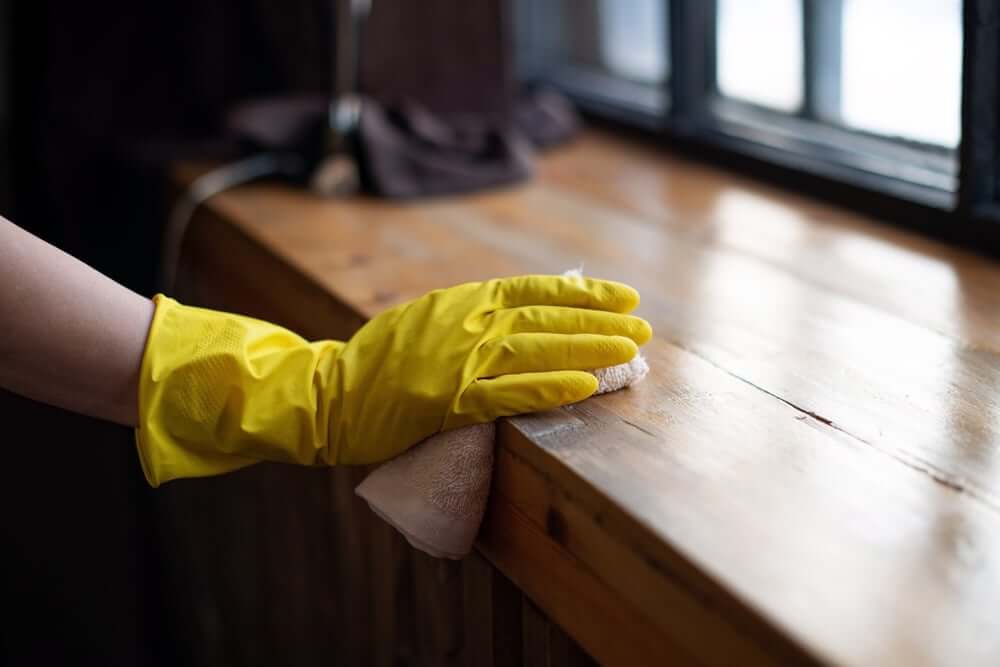 A gloved hand carefully wiping down a wooden windowsill with a cloth, ensuring all dust is removed after the floor sanding and refinishing process by Mr Sander®. The windowsill, like the newly refinished maple hardwood floor below, showcases a smooth, polished finish. The use of yellow gloves highlights the attention to cleanliness and detail in the post-sanding cleanup process.