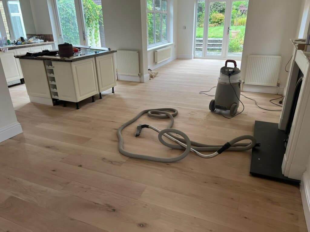 Spacious kitchen and living area with newly refinished wooden floors by Mr Sander®. The room features large windows overlooking a garden and a central kitchen island. A vacuum cleaner is placed on the floor, indicating a recent deep cleaning.
