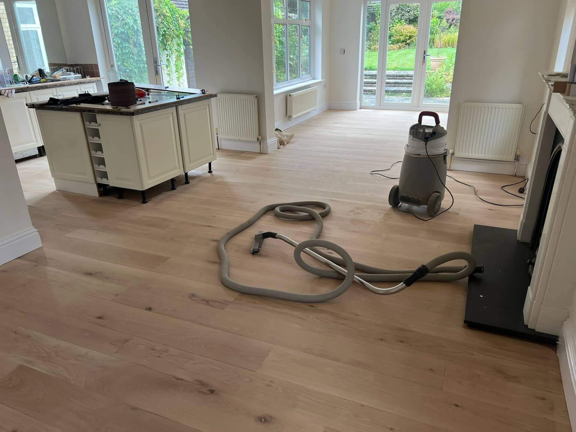 A kitchen and living area with light wooden floors that have been recently sanded. A vacuum cleaner with a hose is placed on the floor, indicating ongoing cleaning or preparation work. The room is bright, with large windows and glass doors leading to a garden. The kitchen island and some tools are visible, suggesting that the refinishing process is in progress.