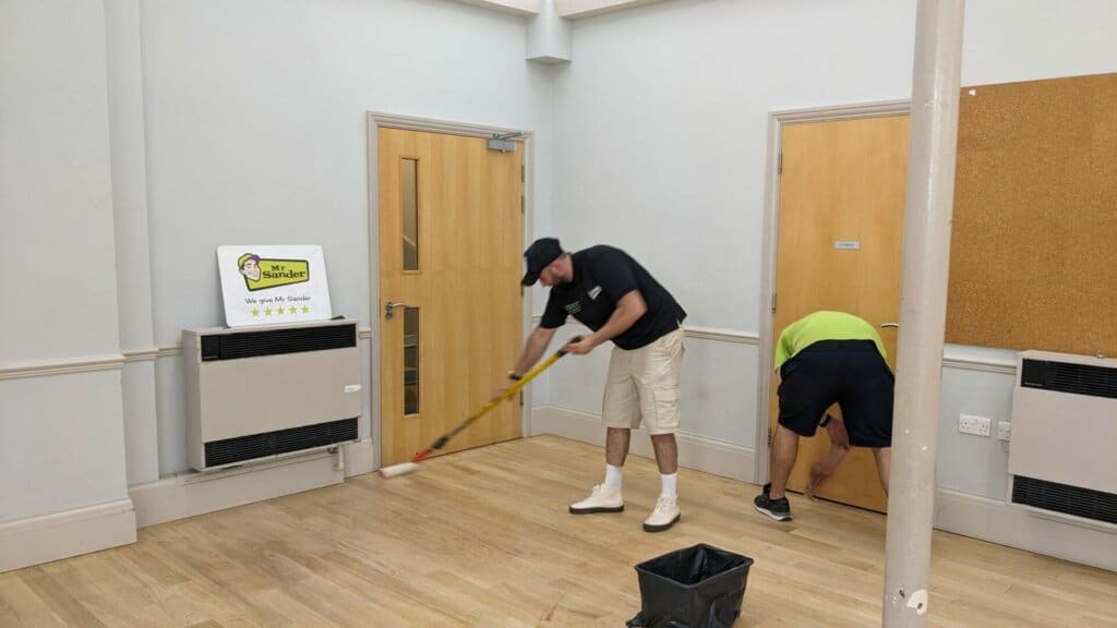 Two workers from Mr Sander® are staining and finishing a wooden floor in a large room. One worker, dressed in a black cap, black shirt, and white shorts, applies a finish with a mop, while the other worker, in a green shirt and dark shorts, focuses on the edges. A sign with Mr Sander's logo and a five-star rating is visible in the background.