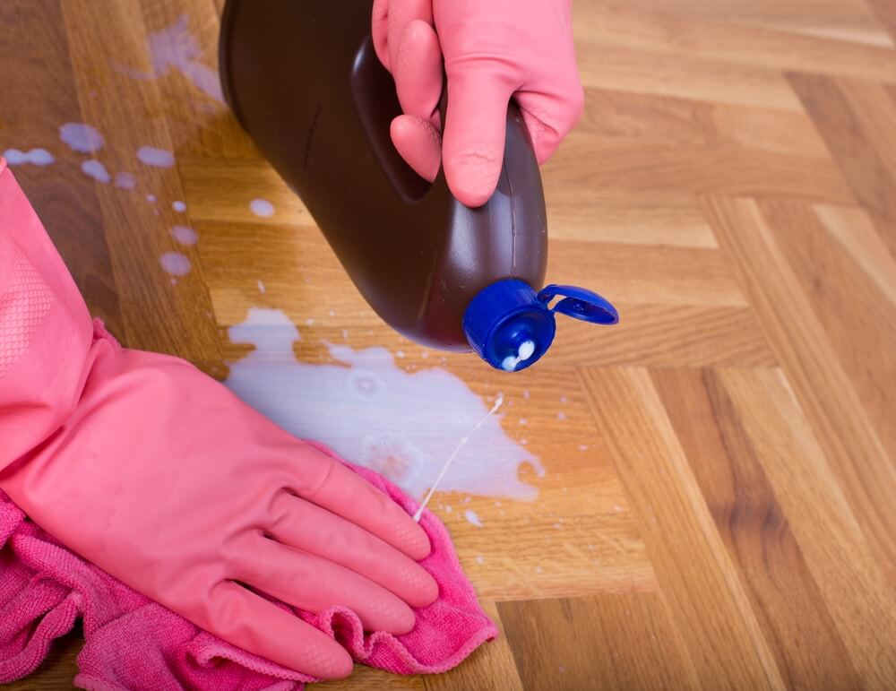 A person wearing pink rubber gloves is cleaning a wood parquet floor. They are pouring liquid cleaning solution from a bottle onto the floor, with a pink cloth used to wipe up the excess. The parquet floor has a polished finish, and the image focuses on the cleaning process to maintain the floor's shine and appearance.