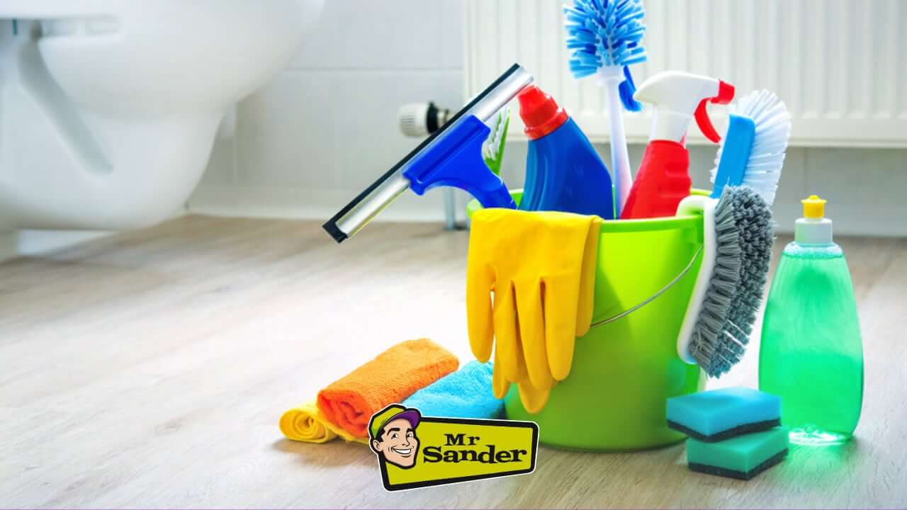A collection of cleaning supplies in a green bucket, including gloves, brushes, sponges, and cleaning liquids, placed on a wooden floor with the Mr Sander® logo.
