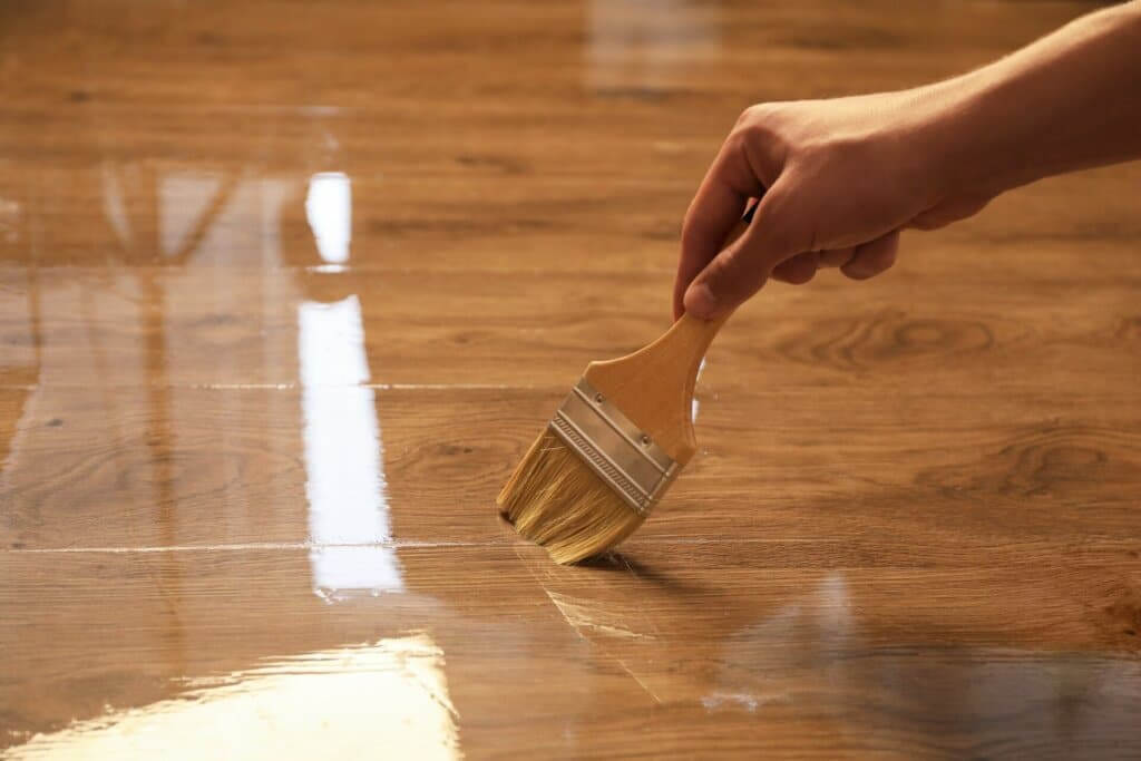 A hand holding a brush applies a clear finish to a wooden floor, creating a glossy and polished surface. The floorboards have a natural wood grain, and the reflection of light is visible on the newly coated section. The image captures the precise application of the finish, highlighting the quality of craftsmanship.