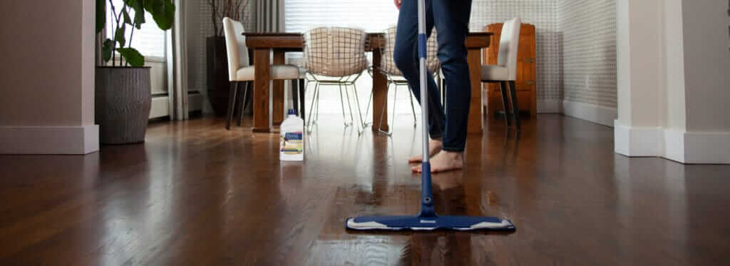 A person cleaning a dark oak hardwood floor with a mop in a modern dining room setting, with cleaning products visible in the background.