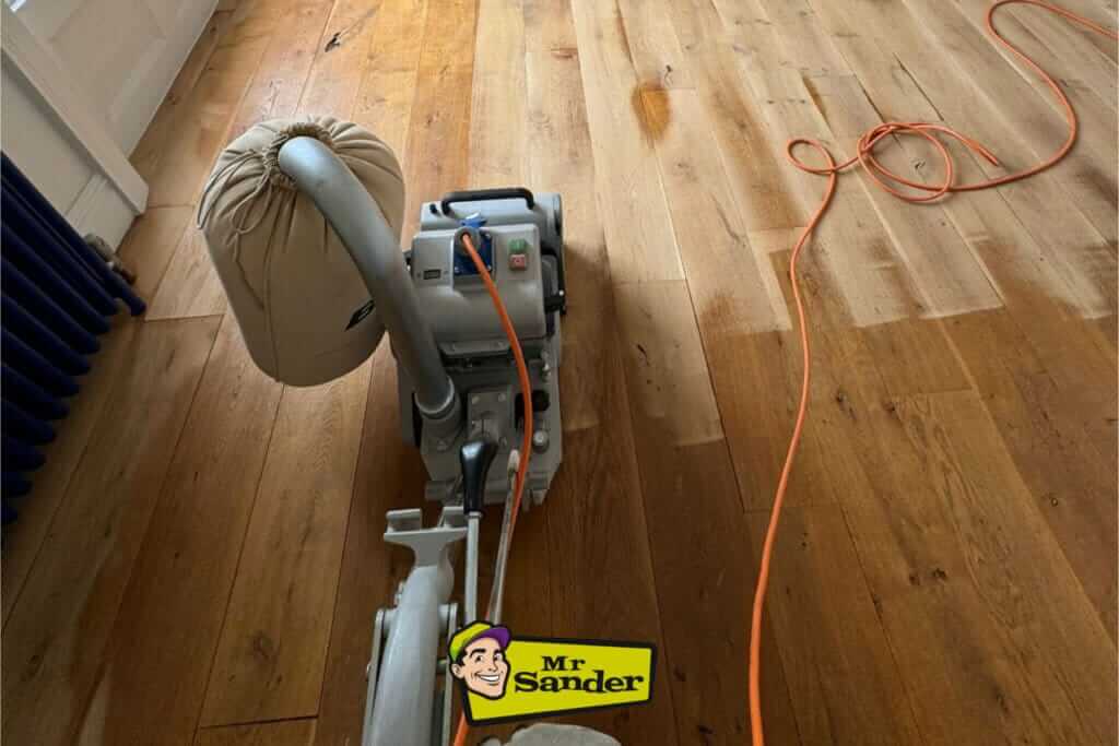 A professional from Mr. Sander® is sanding a parquet floor in a living area in the UK using a Bona Belt sander (AMO110000.3). The machine features a 2.2 kW motor, 230 V voltage, a drum width of 200–250 mm, and a sanding belt size of 200x750 mm.