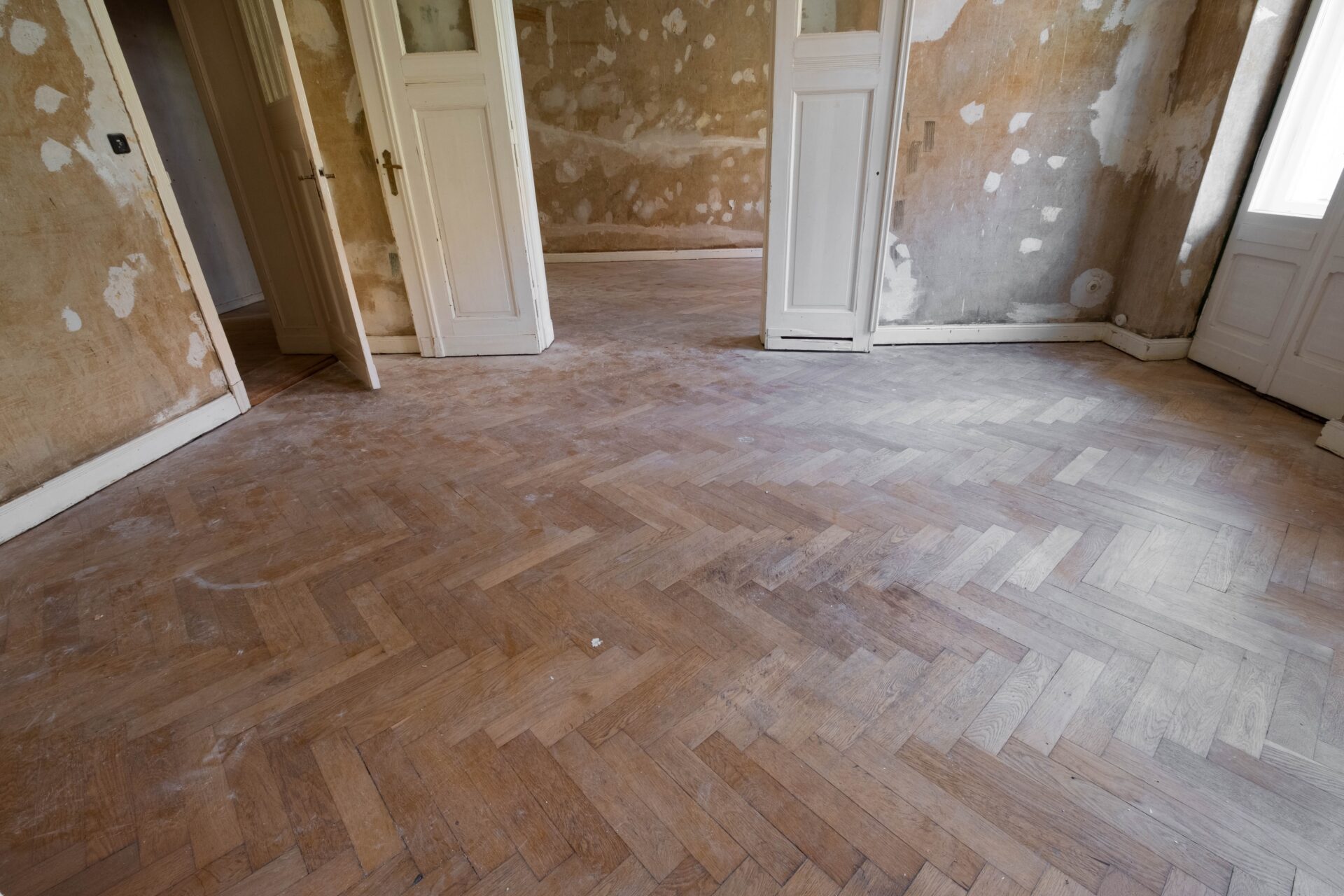 A wide-angle view of a room undergoing wood floor repairs on a dusty, vintage oak herringbone parquet floor before restoration.