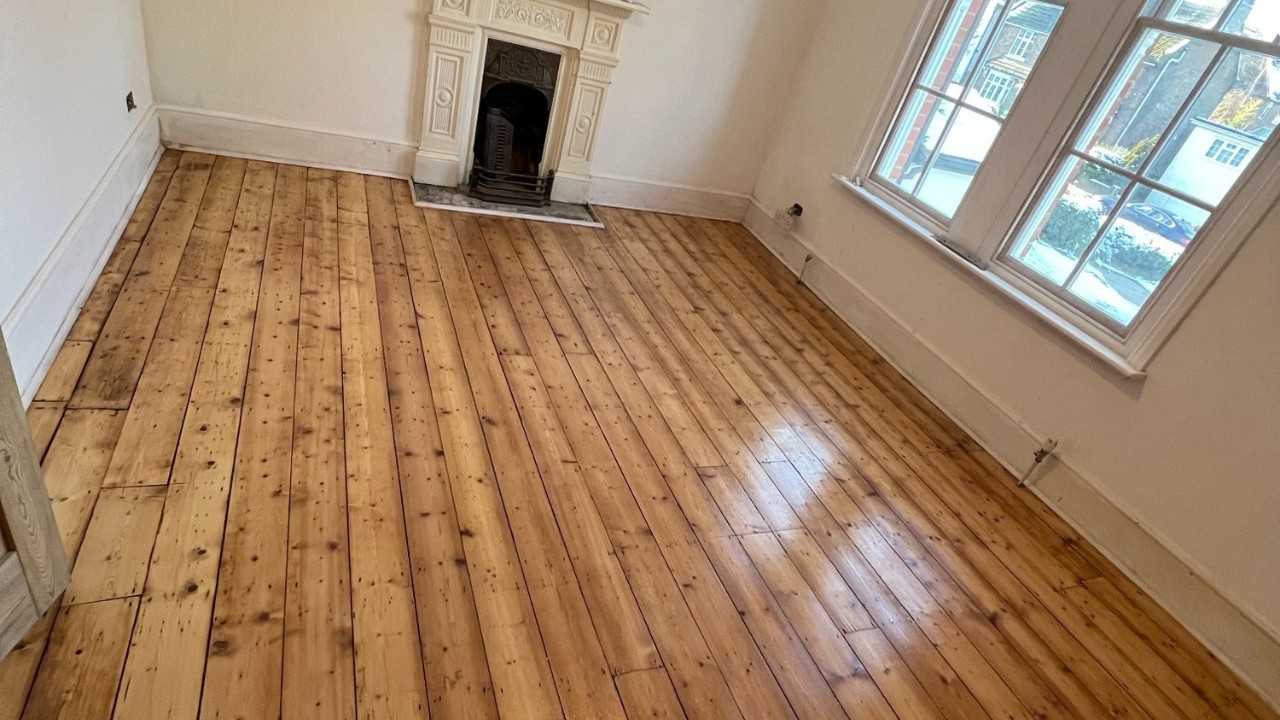 Completed Wood Floor Restoration showing restored pine floorboards with a smooth, sealed finish in a bright living room with bay windows and fireplace.