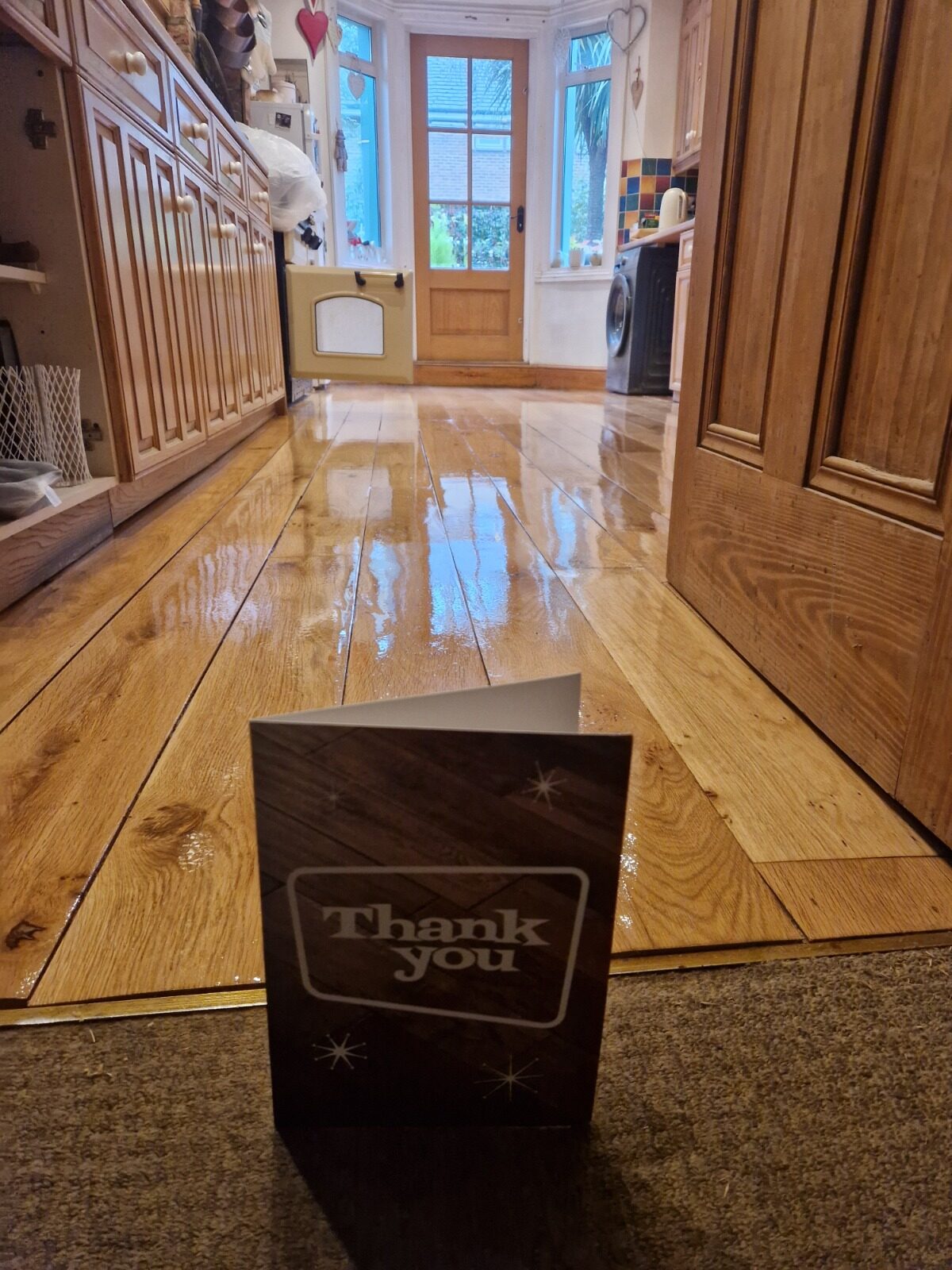 A professionally finished solid pine floor in a kitchen area showing high-gloss results after wood floor sanding, with a "Thank You" card in the foreground.