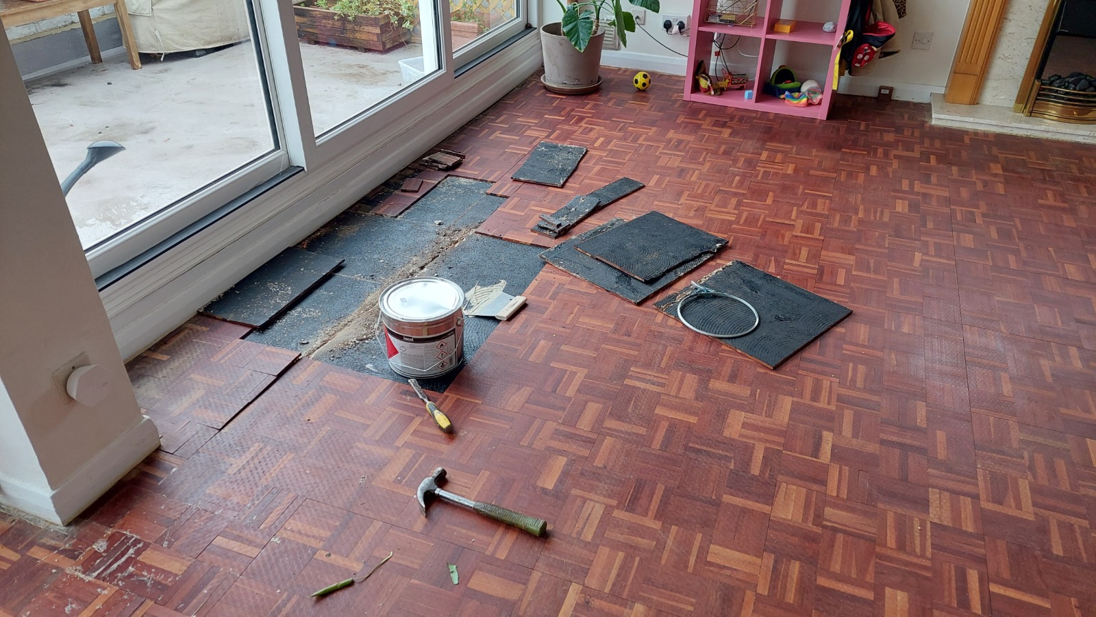 Preparation for Wood Floor Sanding in London on a dark-stained mosaic parquet wood floor, with lifted parquet blocks, adhesive sheets, tools and repair materials near large patio doors.