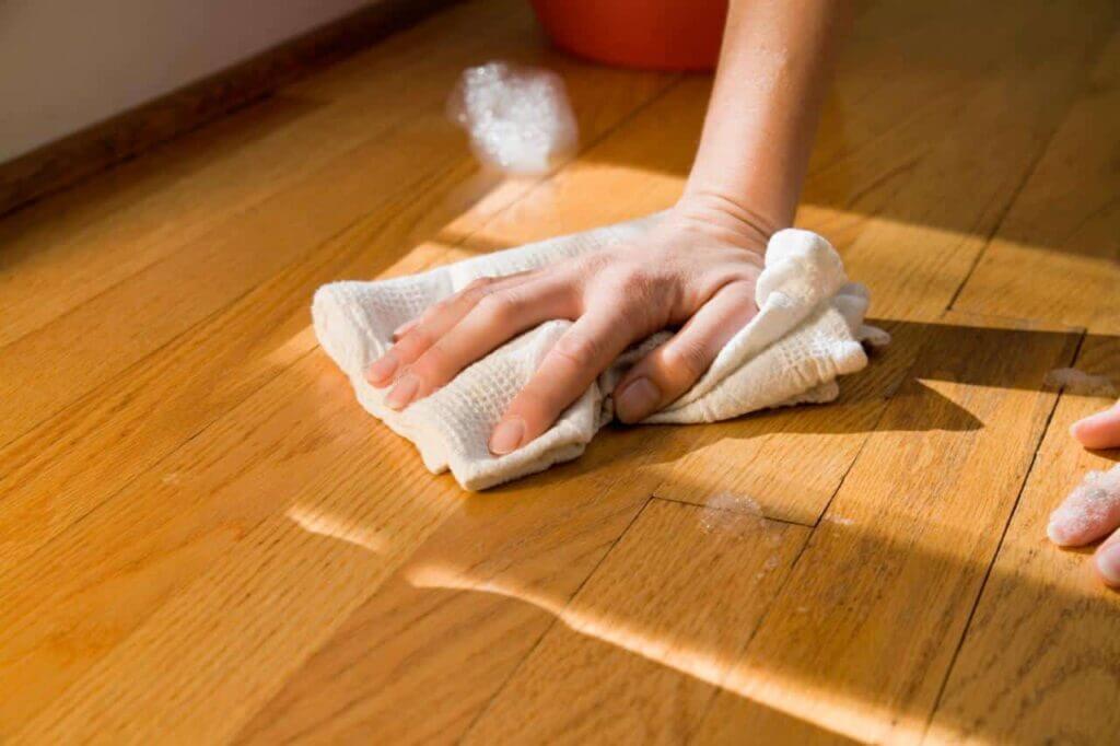 A close-up view of a hand wiping a spill on a maple hardwood floor using a white cloth. The floor showcases its natural grain and smooth finish, reflecting light from the nearby window. A small bucket is visible in the background, indicating the cleaning process. This image highlights the importance of maintaining the quality of the refinished floor done by Mr. Sander®.