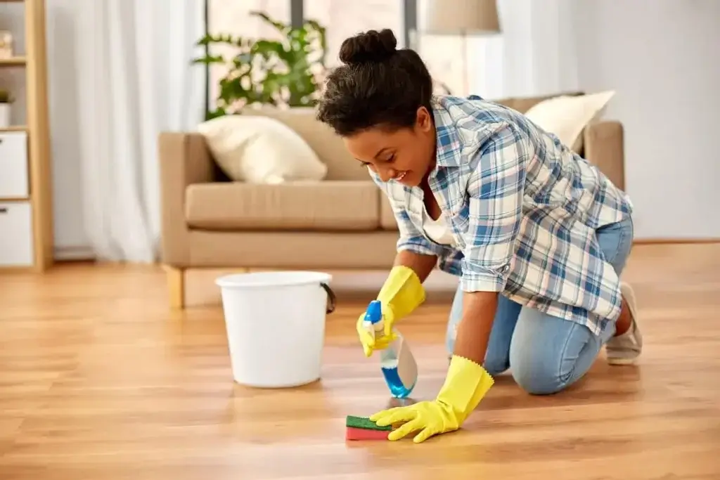 Smiling woman kneeling on a newly refinished wooden floor by Mr Sander®, wearing yellow gloves and using a sponge and cleaning spray to wipe the floor. A bucket is placed beside her, and a cozy living room with a sofa and plants is in the background.