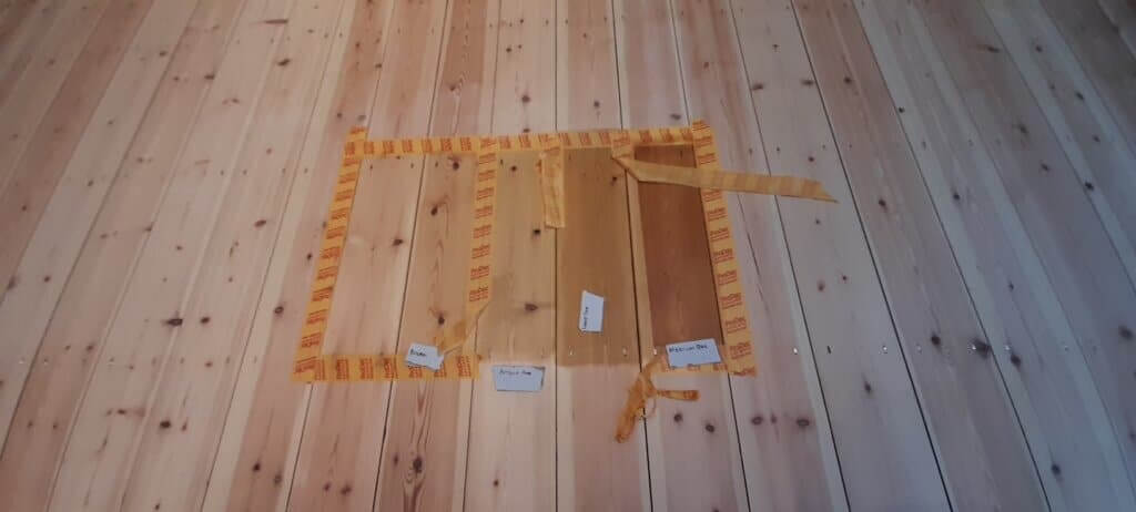 A light wood floor with test patches showing various wood stains and finishes, bordered with yellow tape for comparison.