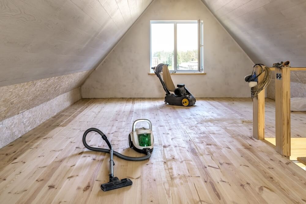 A freshly sanded pine wood floor in an attic room is being prepared for refinishing. A vacuum cleaner and floor sander are placed on the smooth surface, ready for dust removal before the final finish is applied. The room has slanted walls and a large window providing natural light.