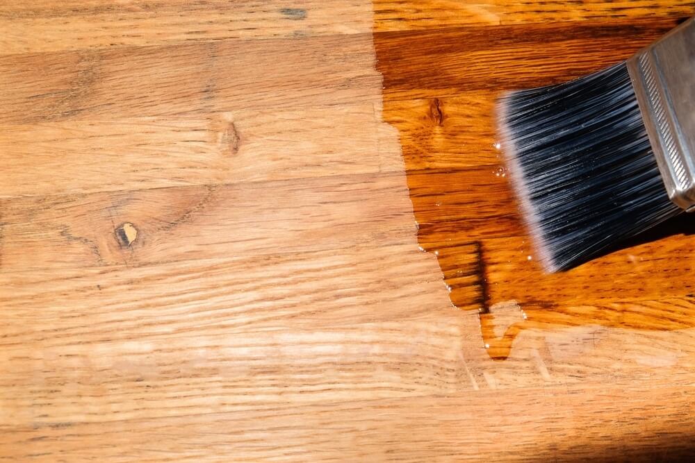 A close-up view of a paintbrush applying a clear finish or stain to a wooden floor. The wood grain is visible, with the finish enhancing the natural color and texture. The contrast between the treated and untreated areas shows the difference in color and sheen.